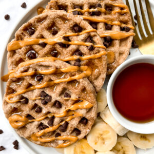 top view of waffles drizzled with peanut butter next to banana slices and ramekin of syrup
