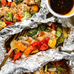 Chicken foil packets on a baking sheet next to a dish with teriyaki glaze