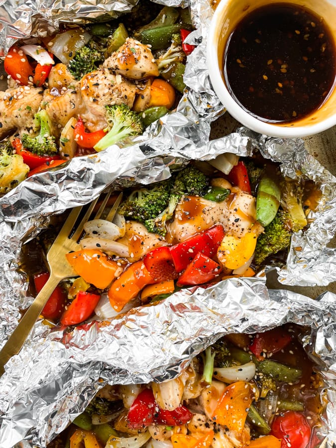 Chicken foil packets on a baking sheet next to a dish with teriyaki glaze